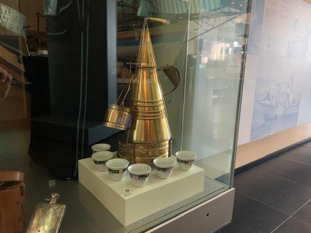 A shiny brass coffee pot that is shaped like an upside-down cone. It is surrounded by 6 small cups.