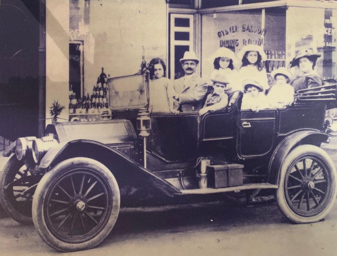 A black and white photos showing an old fashioned, open topped car outisde a building. The window of the building is labelled “Oyster Saloon Dining Room’. Inside the car are an adult male and female in old fashioned clothes, with 6 small children.