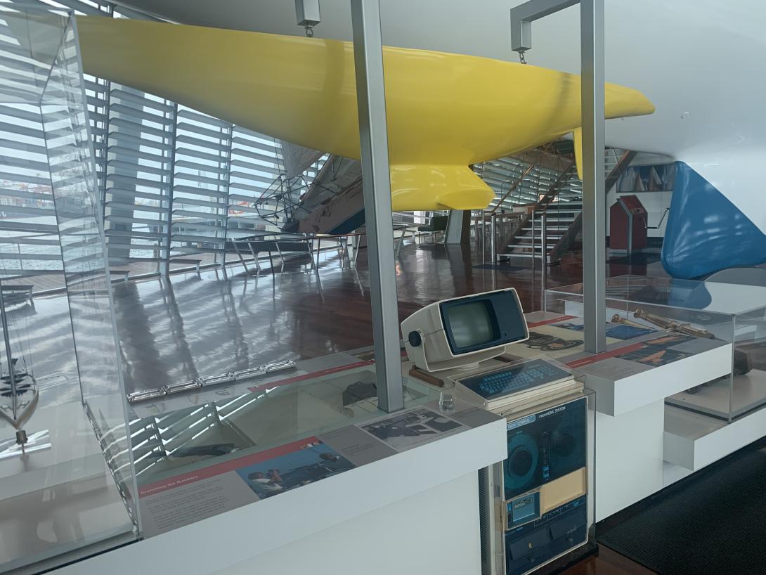 A small-scale yellow yacht hull is suspended above a display of an old 1980s computer.