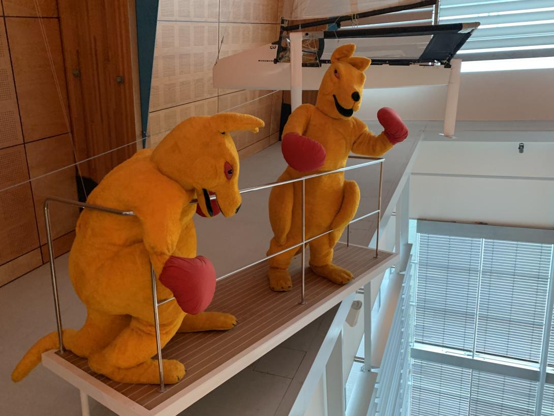 Two yellow Kangaroo mascots wearing red boxing gloves stand on a platform overlooking the museum gallery below.