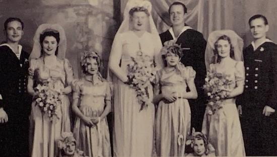 Three pairs of brides and grooms stand together, with 4 flower girls in attendance. All brides wearing wedding dresses and carry bouquets of flowers and the grooms wear naval uniforms.