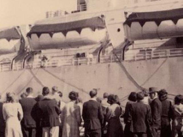 A black and white image of a ship docked at port, with people dressed in 1940s clothes standing on the dock.