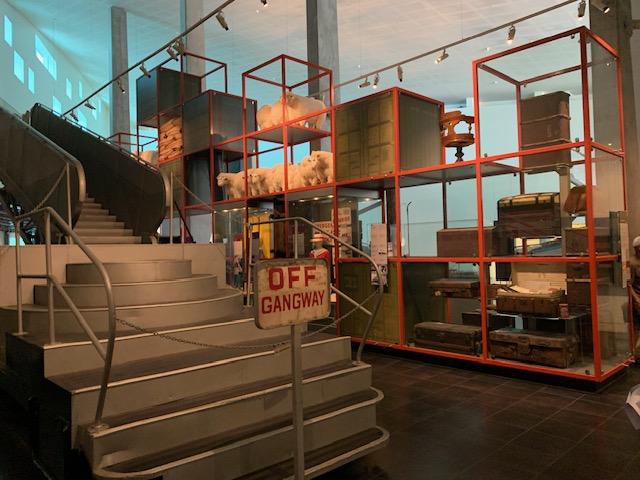 Alt text: A metal ship’s gangway is displayed with a metal sign stating ‘OFF GANGWAY’. Behind is a tall display featuring suitcases and packing crates.