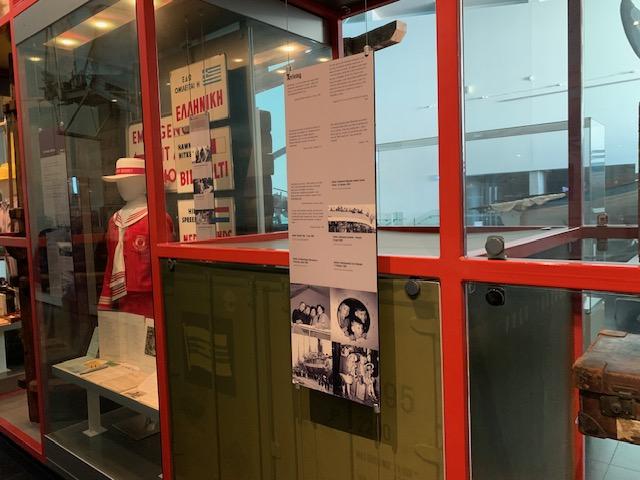 Alt text: A sign with text and black and white photos hangs in front of red metal exhibition framework. In the background is a mannequin wearing a red uniform and white hat.