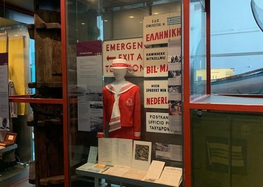 ALT TEXT: A female mannequin wears a red jacket with a white and red striped collar and cuffs, and a red skirt. It wears a white hat with a red hat band. Behind is a collection of signage in foreign languages.
