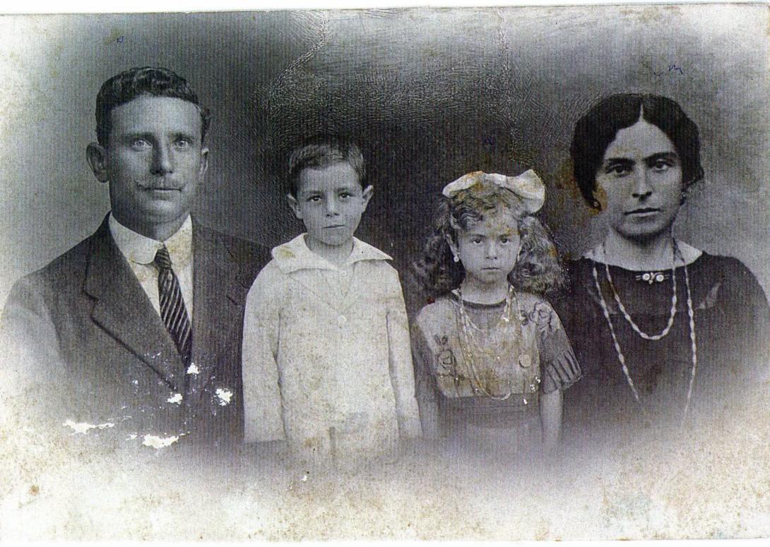 A black and white photo showing a man with a moustache in a suit and tie, a woman with dark hair wearing a double stranded necklace, with two small children (a boy and a girls) between them. He wears a light coloured suit with a large collar, and she has a large bow in her hair and earrings in her ears.