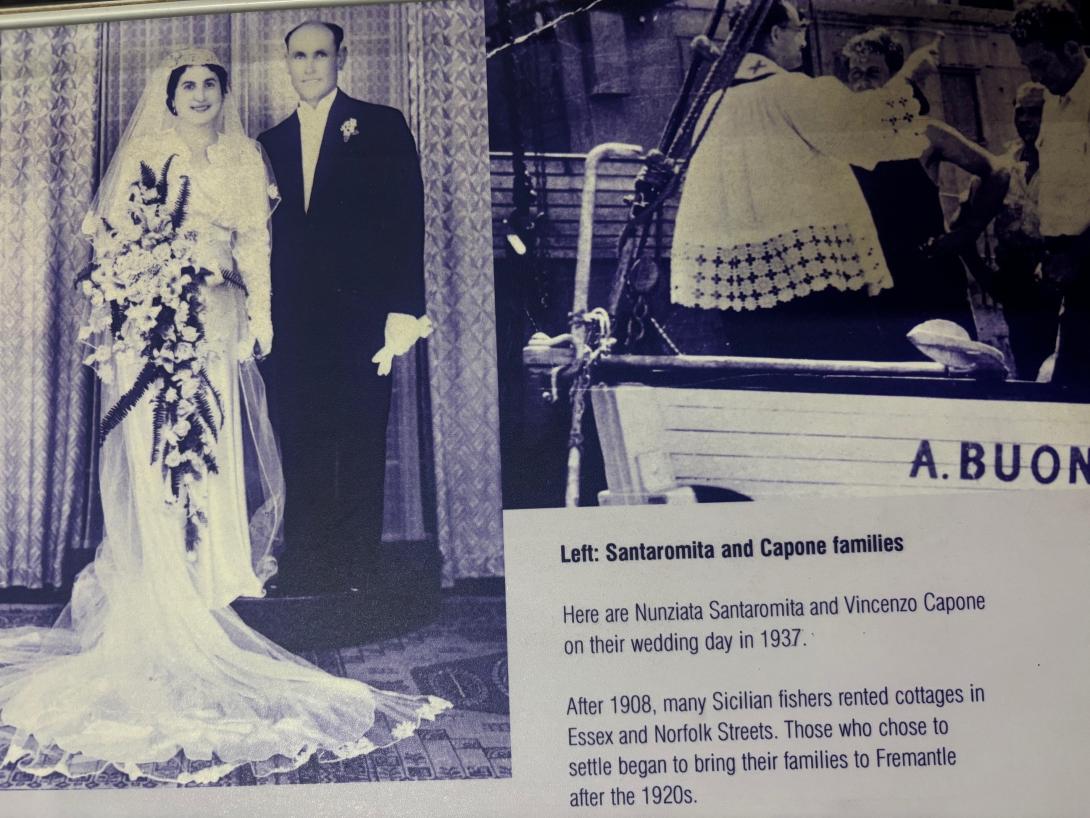 A bride and groom stand for a formal wedding photo. She has a long white train and is carrying a large bouquet of flowers that trails in front of her dress. He wears a dark suit and is holding his gloves