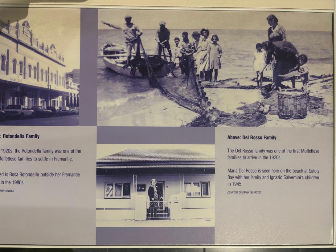 A black and white photo showing a group of adults and children on the shore with a large fishing net and basket. Two men stand on a small boat that is pulled up to the shore.