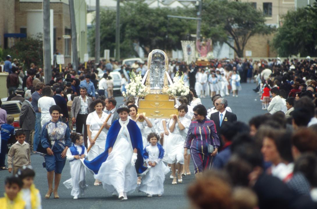 A woman in a long white dress and royal blue cape walks in a parade. She is surrounded by children and other women, and leads a group of women who carry a large statue of the Virgin Mary on their shoulders.