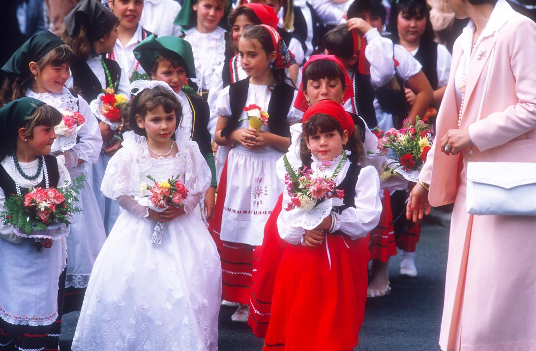 Small children are gathered together to walk in a parade. One wears a white dress and veil that resembles a communion gown. Others wear red or green head scarves with matching skirts, some with white aprons. they carry bouquets of flowers in their hands.