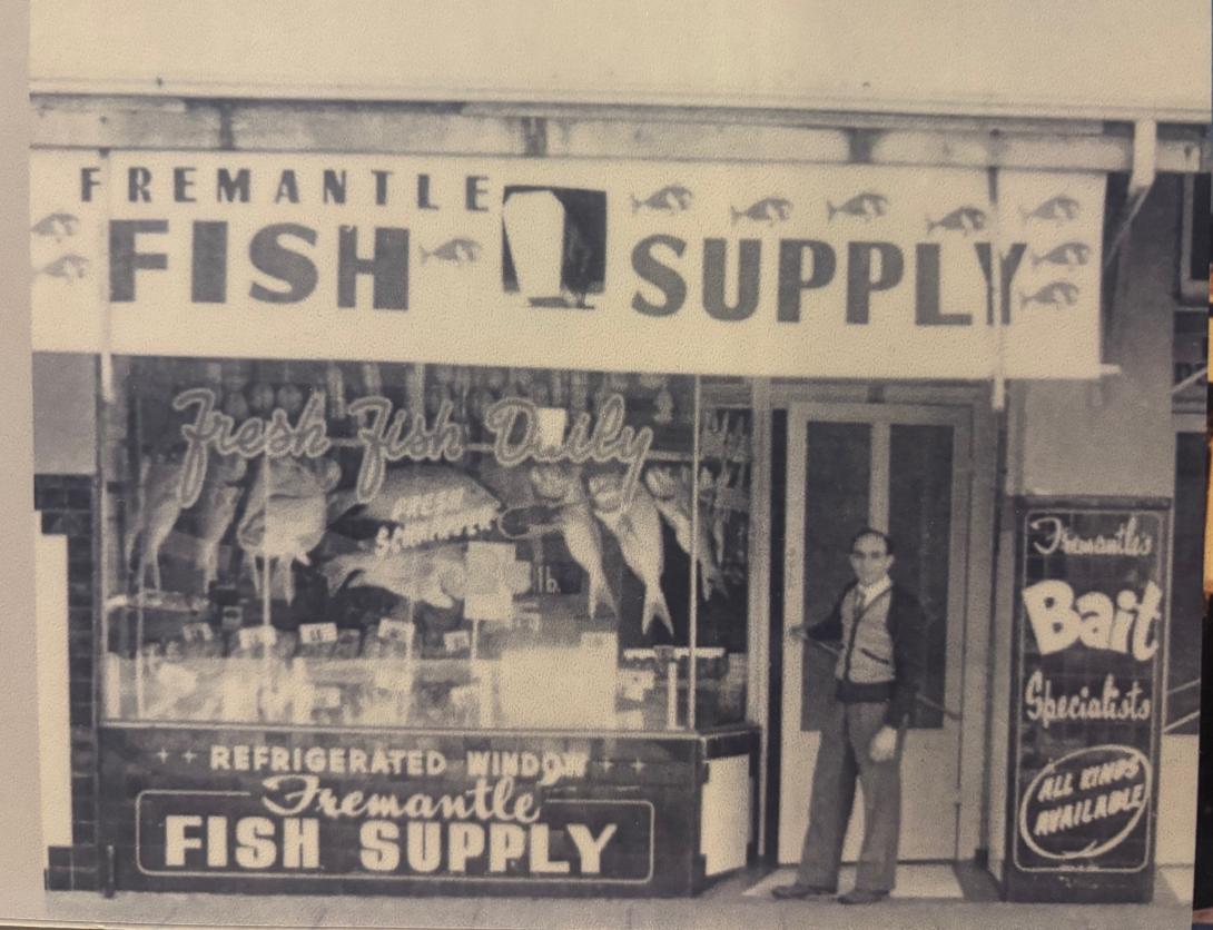 A black and white image showing a vintage fish supply store, with its front window advertised as 'refrigerated' and displaying fresh fish. A man in neat trousers and a cardigan, shirt and tie stands at the front door with a welcoming smile on his face.