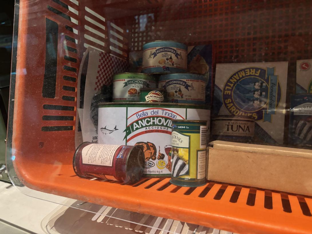 An orange plastic basket lies on its side, and contains an assortment of different size cans of anchovies, tuna and sardines