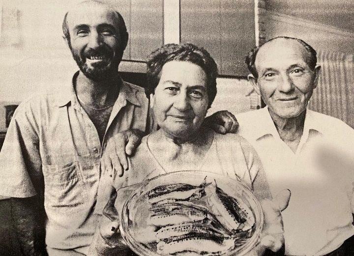 A smiling older lady displays a dish of frsh sardines. she stands with her grown son on one side and an older man on her other.