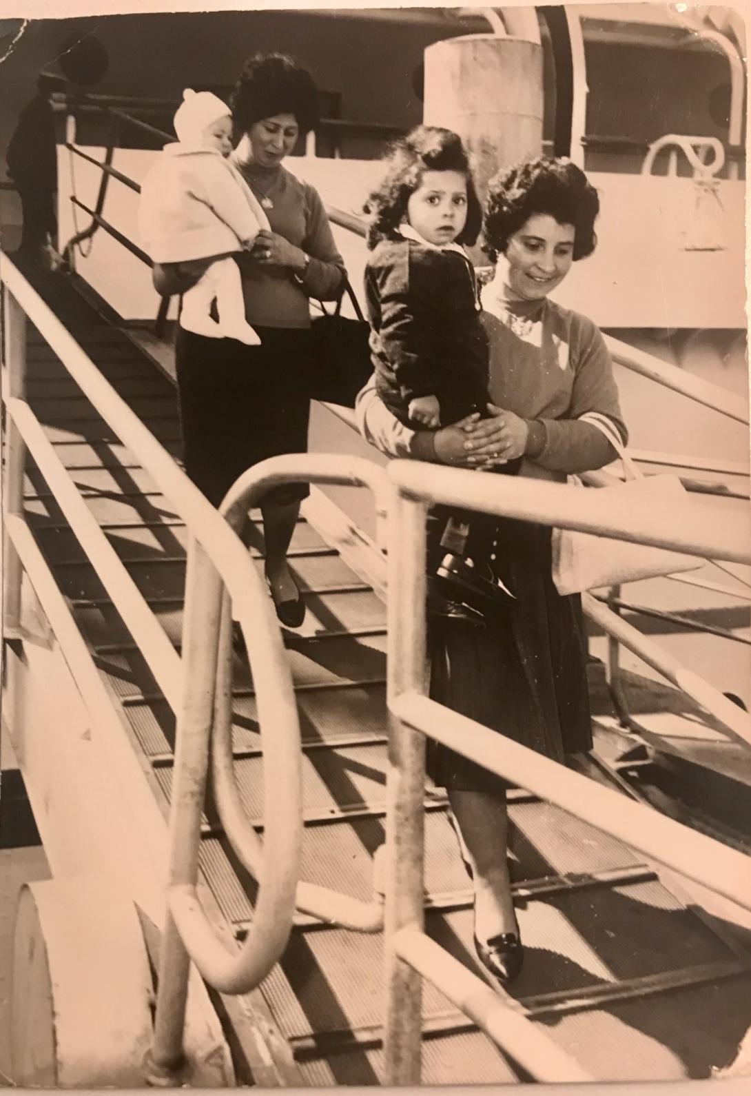 Two dark haired women, each holding a small child, disembark from a ship down a gangway.