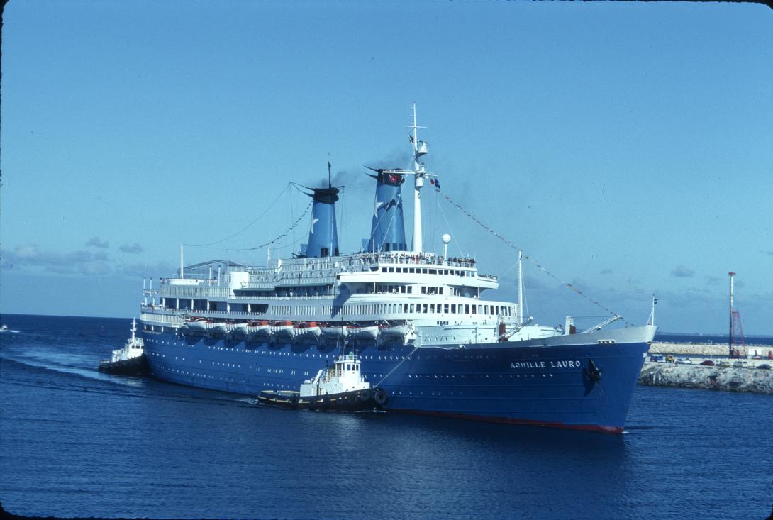 A cruise liner with a blue hull and white upper sails into port on smooth waters. Tug boats guide it in, and the sky and water are both bright blue