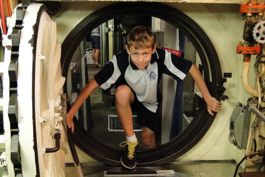 A boy in school uniform climbs through a narrow submarine hatch, a cheeky grin on his face.