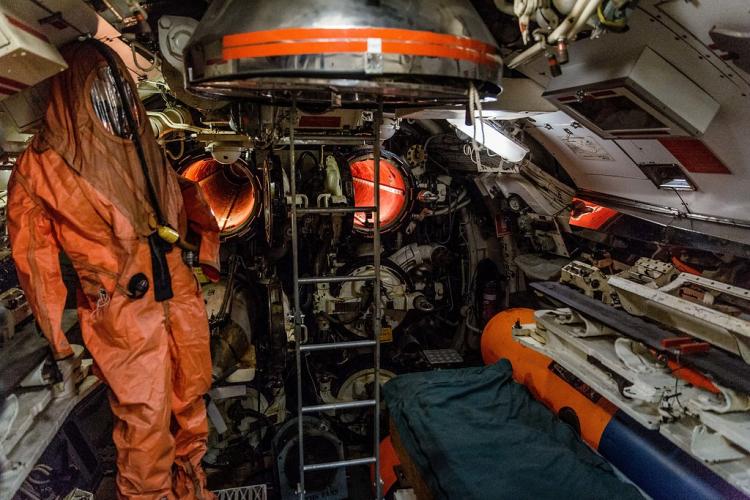 An orange protective suit with full face shield hangs next to four torpedo tubes.
