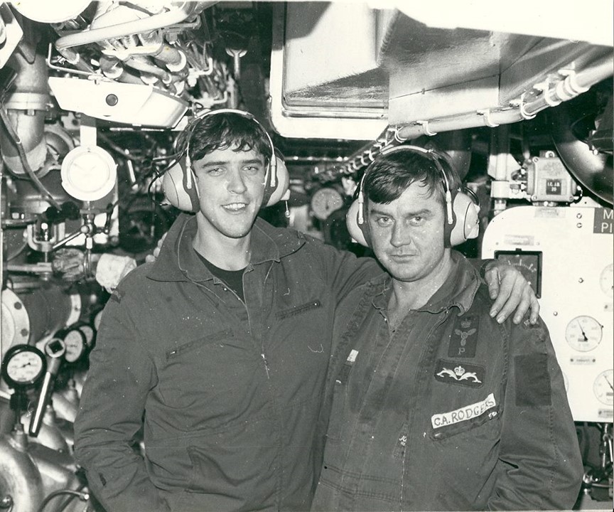Two submarine crew wearing hearing protection smile at the camera inside a cramped compartment. One has his arm around the other's shoulders.
