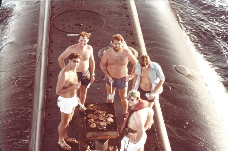 On the top of a submarine in the water stand five submarine crew, four of them dressed in only shorts, surround meat cooking on a barbecue.