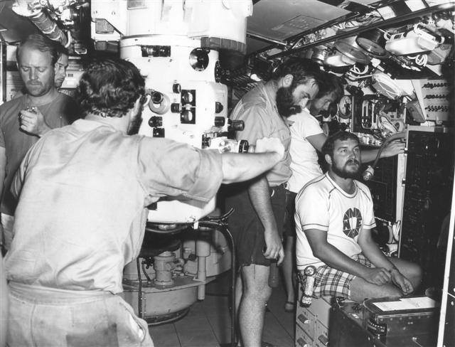 Many submarine crew crowded in a control room. They are all wearing shorts and short-sleeved shirts or casual t-shirts. In the centre is a large periscope with a crew member looking into the eyepiece.