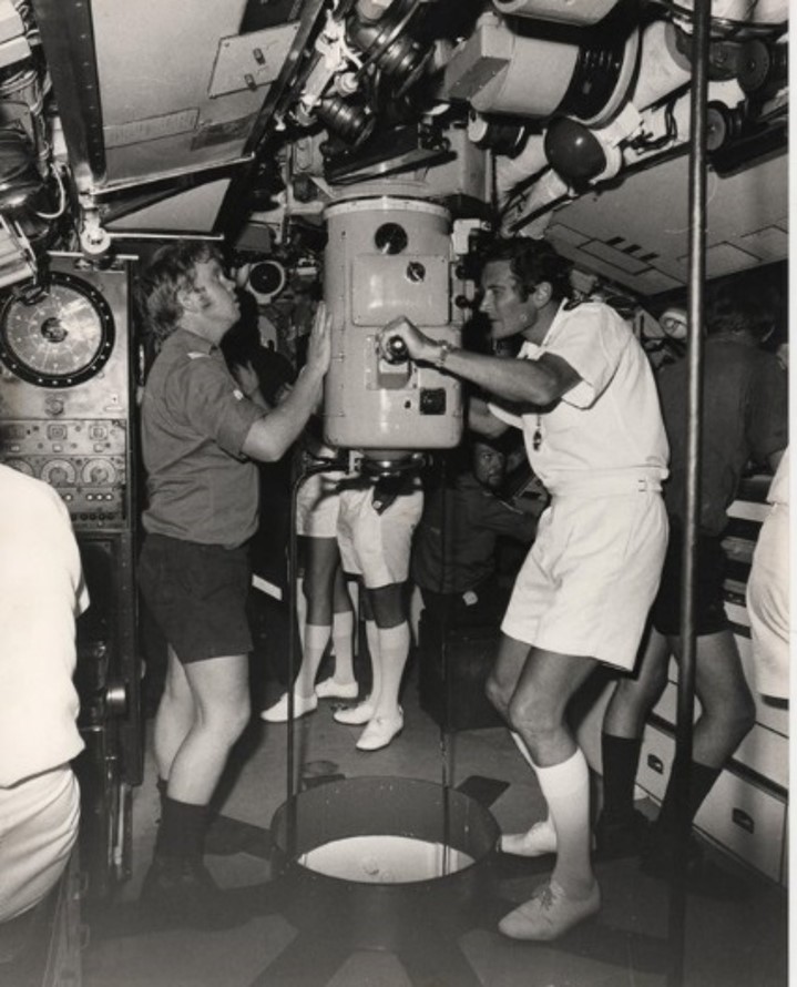 Two submarine crew dressed for a hot environment either side of a periscope. One is looking into the eyepiece.