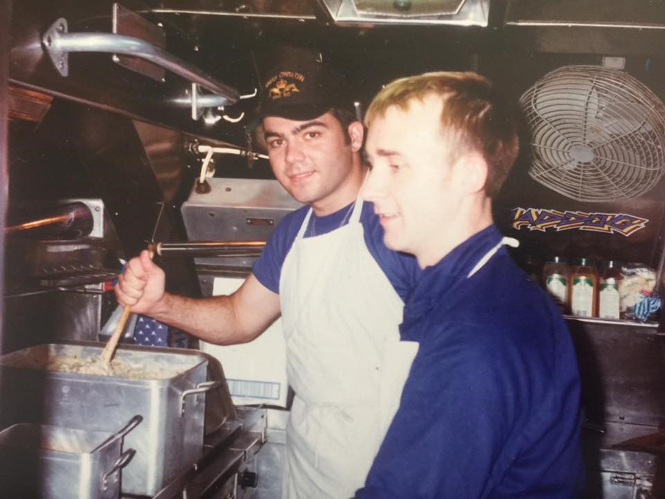 Two young men in blue shirts and white aprons cook at a submarine kitchen. One is wearing a cap with their vessel's insignia.