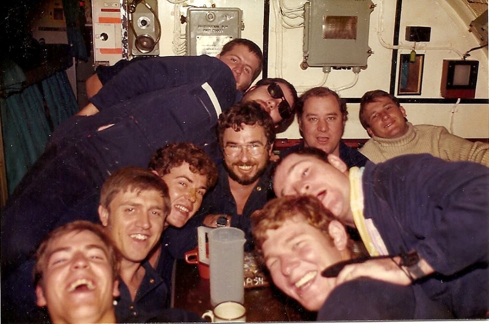 Several submarine crew members crowd around a cramped cabin table smiling at the camera.