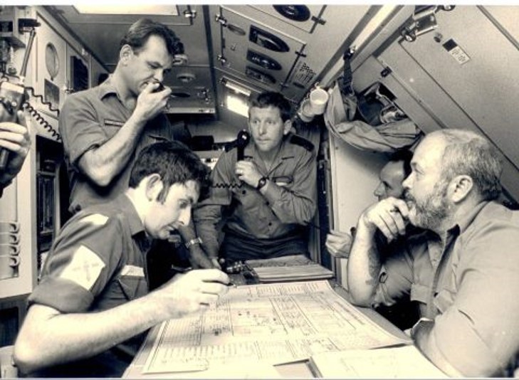Submarine crew gathered around a table looking at charts. One is holding a telephone and another is speaking into a radio.