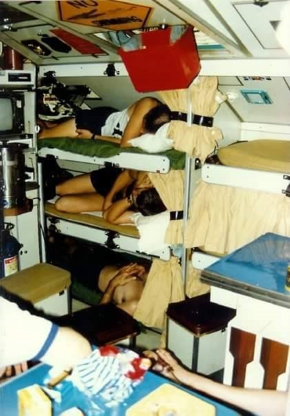 Three bunks stacked vertically against the submarine wall, each with a crew member sleeping. Above the bunks on the ceiling, someone has jokingly mounted a yellow "No Swimming" warning sign with the open mouth of a crocodile.