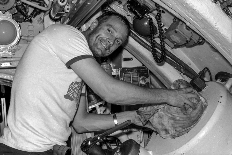 A smiling crewmember in a t-shirt forces a tightly-wrapped bundle of garbage into a narrow metal chute.