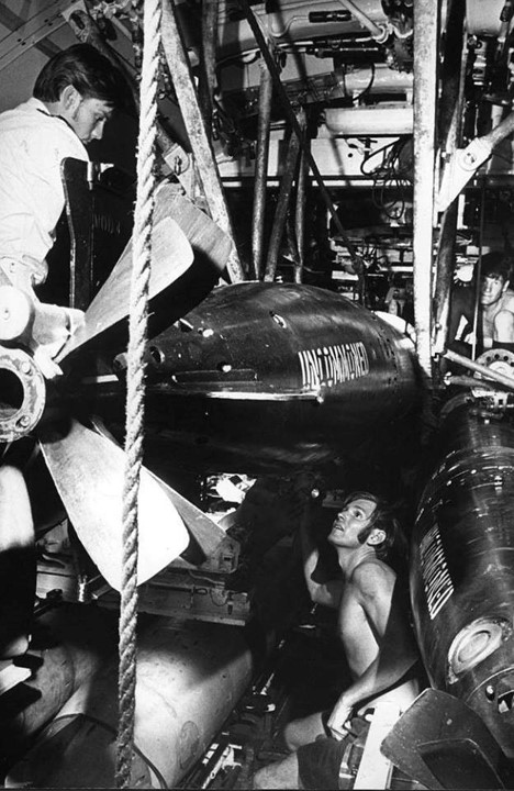A shirtless man is crouched underneath a very large and heavy torpedo as it is loaded into a tube.