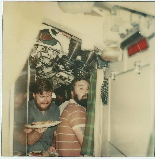 A crewmember with two plates of food squeezes past another in a narrow corridor.