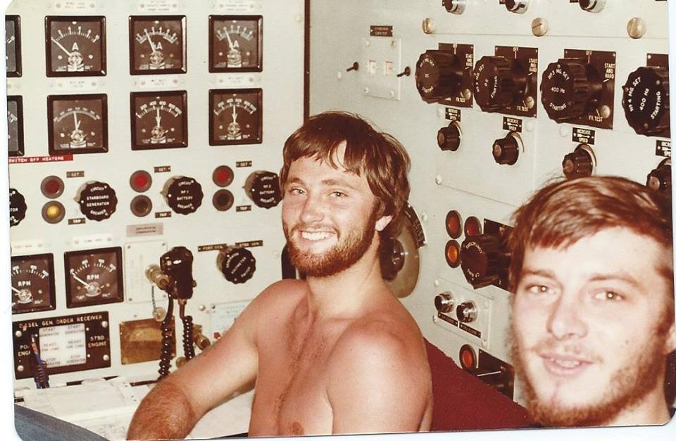 A grinning shirtless submarine crewmember sits in front of a panel of many dials.
