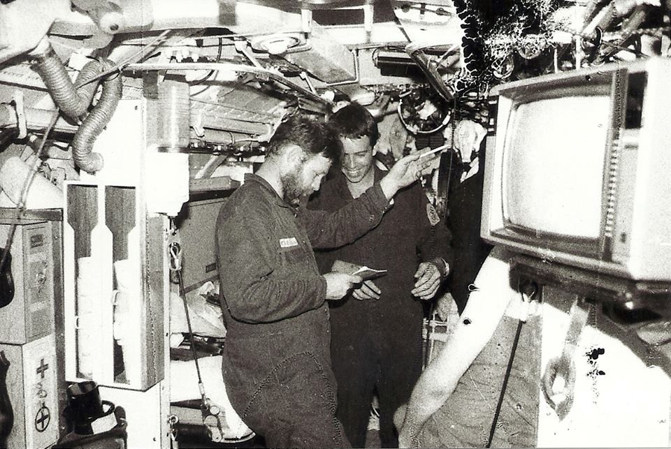 A grainy black and white photo of two crew in overalls working in the tight corridor of the submarine.