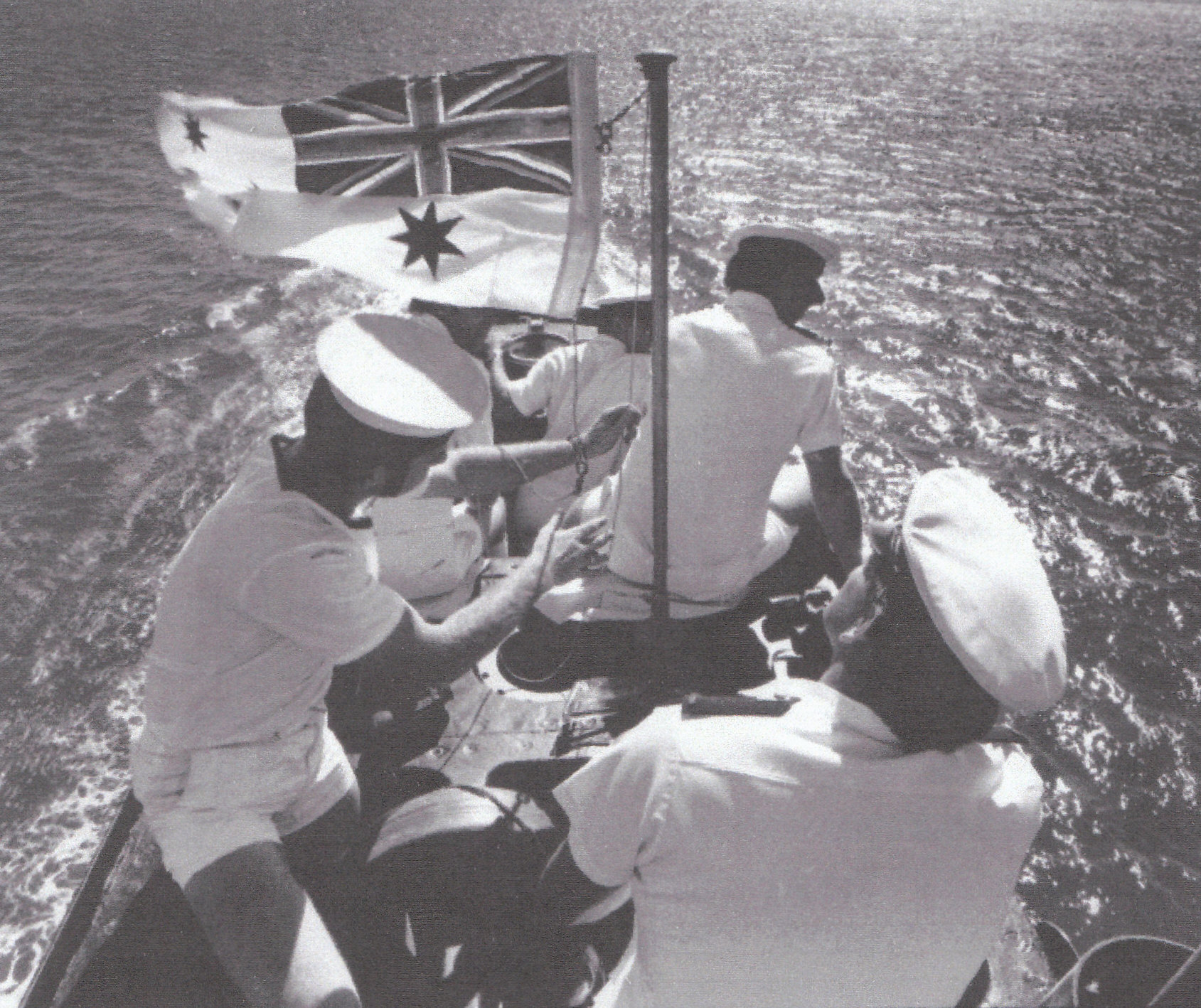 A black and white photo of several sailors in clean white uniforms gathered on top of the sub. An Australian flag is raised.