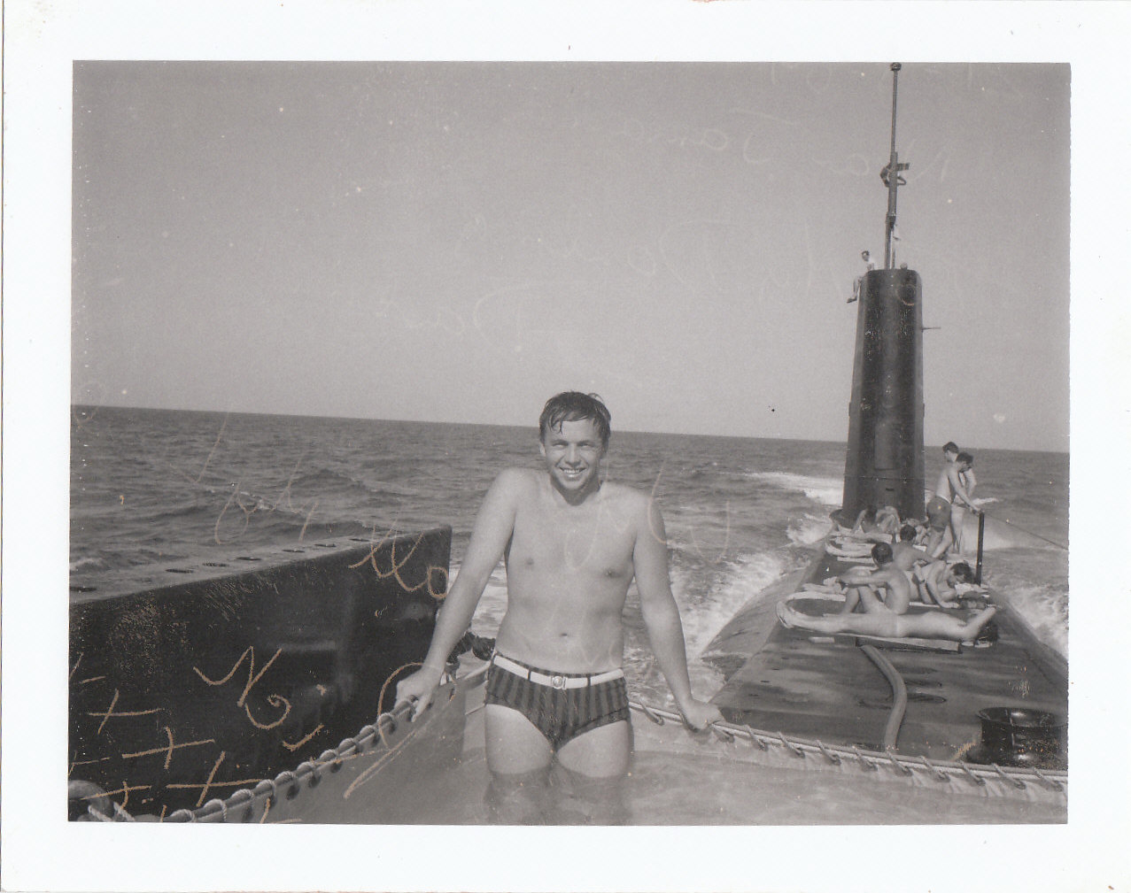 On the top of the submarine, a sailor in a bathing suit smiles from a fabric shallow pool filled with water. Several other men sun themselves on the deck.