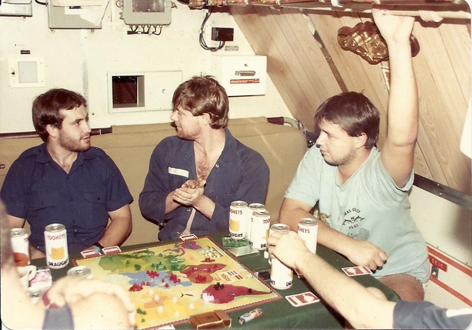 Around a table with many Tooheys Draught beer cans, game cards and a large colourful board game board sit crew members planning their next move in the game.