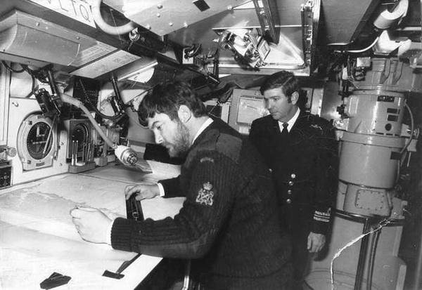 An officer in a dark Navy jacket and tie looks over a crew member working on a nautical chart.