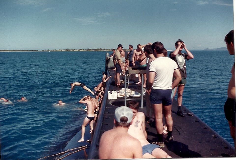 Many crew are gathered in bathing wear on top of the submarine. Some are swimming in the water.
