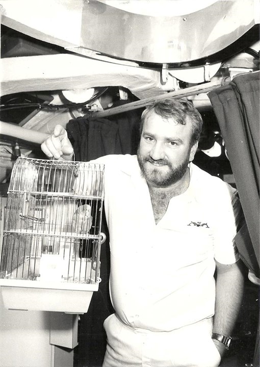 A grinning crewmember holding a small bird cage containing a budgerigar sitting on a perch.