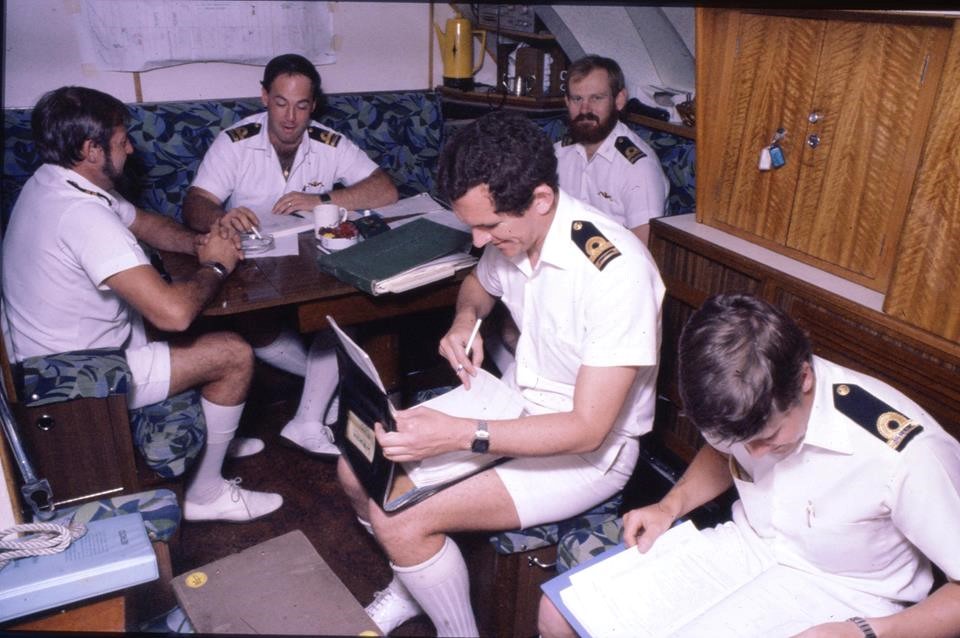 Crewmembers in clean white uniforms lounge around a mess table. One taps a cigarette on an ashtray.