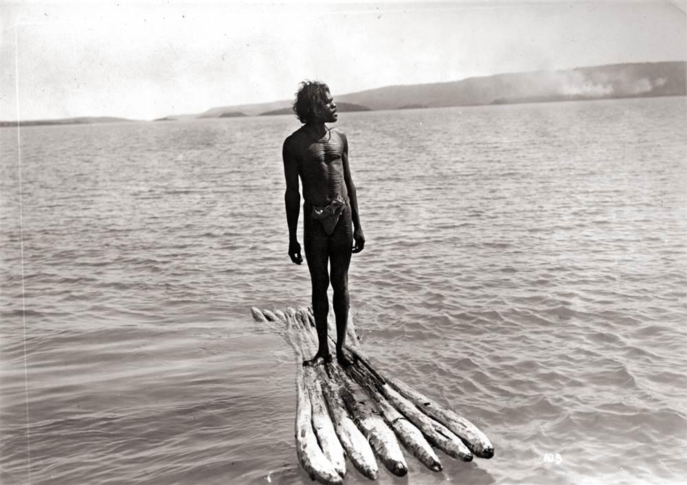 An aboriginal man stands tall on a raft on open water.