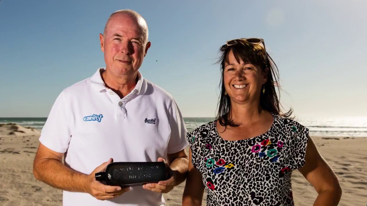 On a beach, a man and a woman smile while holding a dark glass bottle.