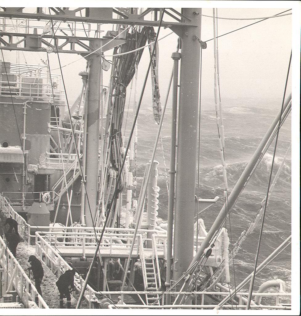 A view from the deck of a ship in rough seas. Crew in heavy clothing are shovelling ice off the deck. There is ice on the railings and other parts of the ship.