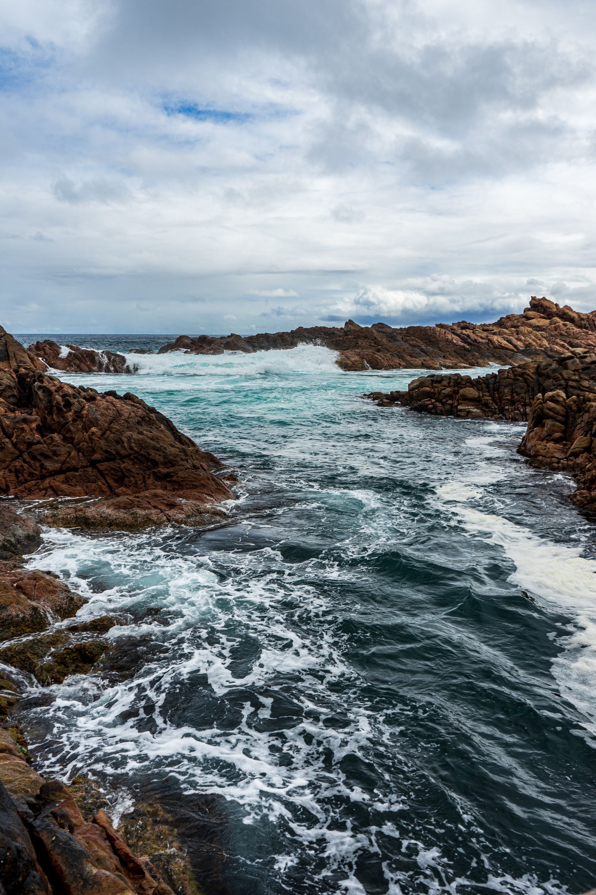 Waves crash against rocks either side of a canal. The ocean is visible in the distance.