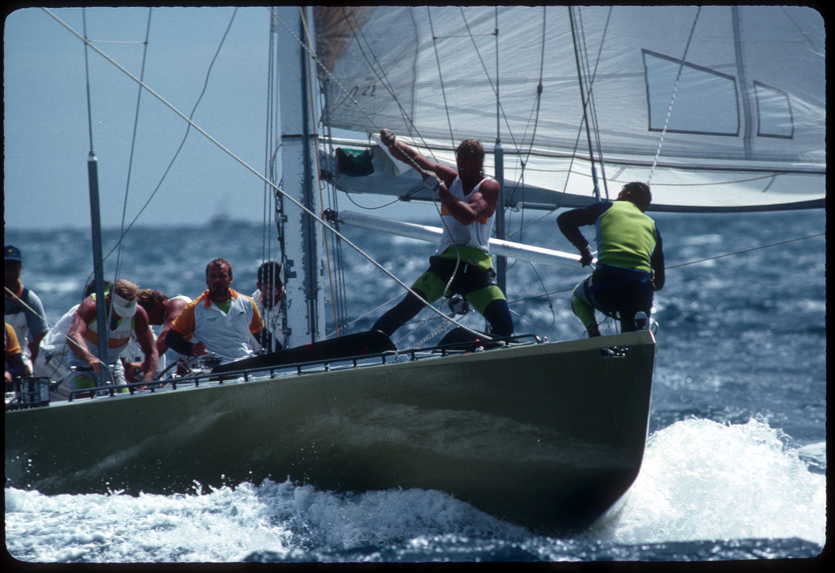 A muscular man in a singlet stands near the bow of a yacht pulling on rigging.