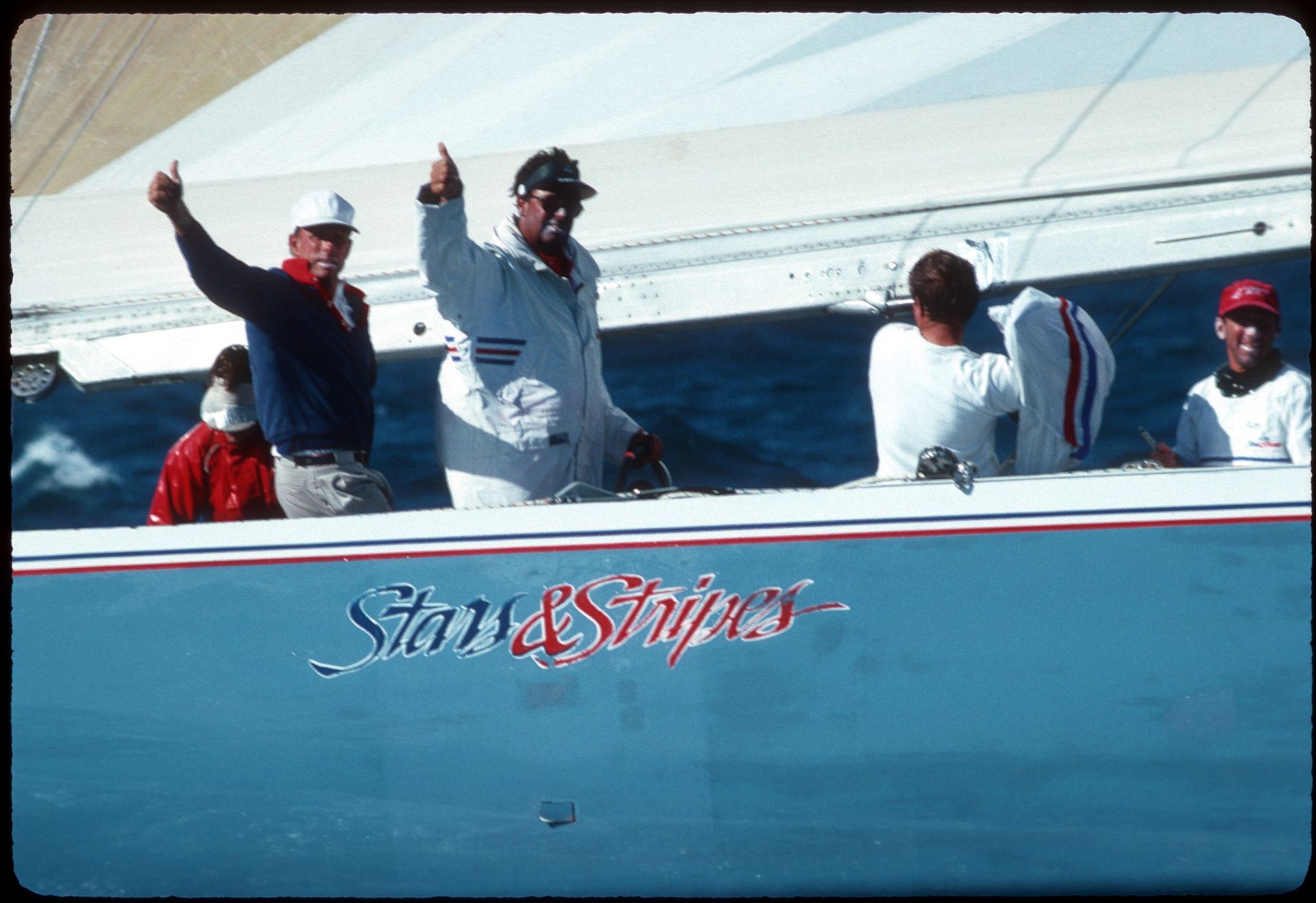 Sailors give a thumbs up to the camera from the deck of a yacht named "Stars & Stripes."