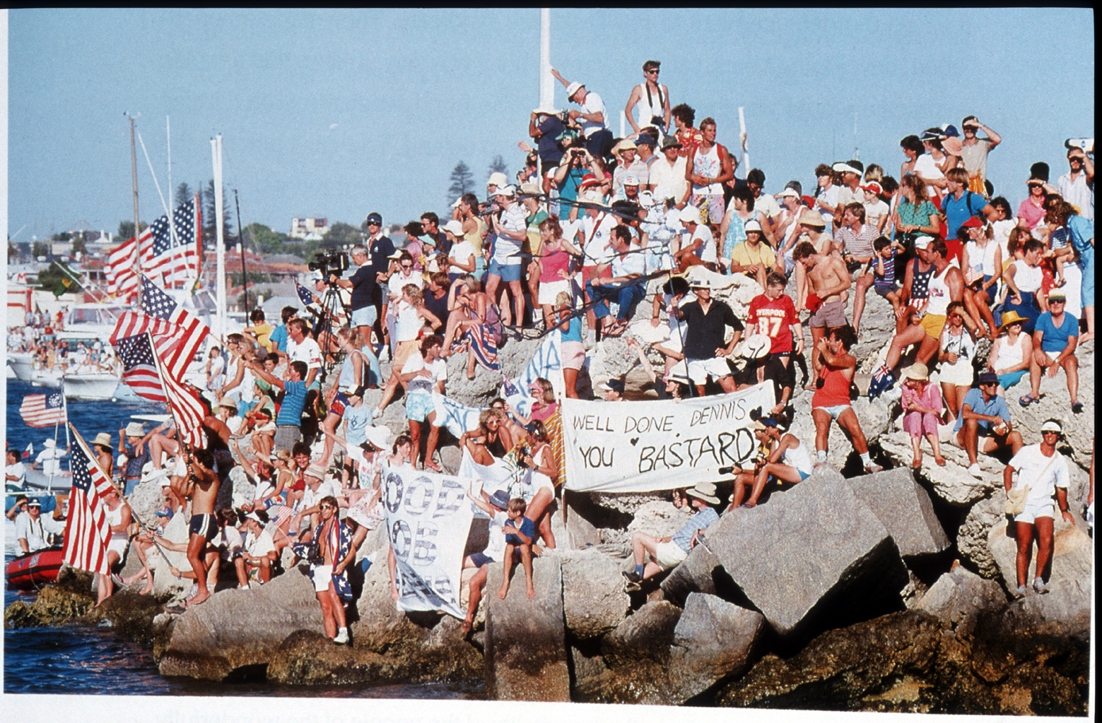 A large crowd almost entirely covers the rocks. A handwritten sign in the centre reads "well done Dennis, you bastard."