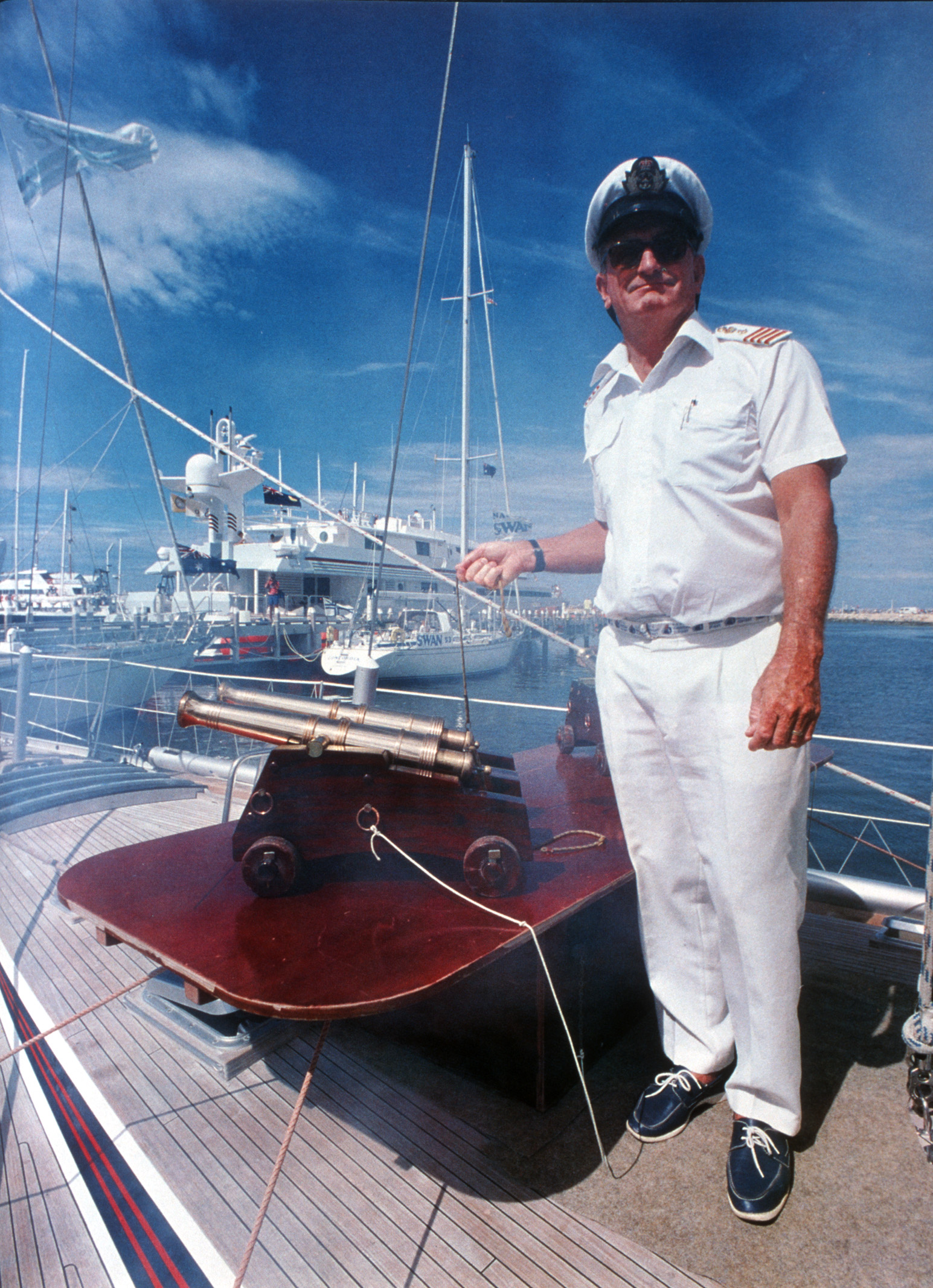 A man in a white sailor's uniform and hat holds the cord of the starting cannon, on the deck of a boat.