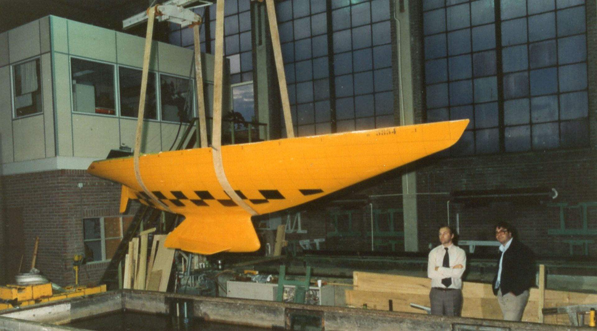 Two men in businesswear observe a large yellow model of the keel suspended in the air above a large water tank.